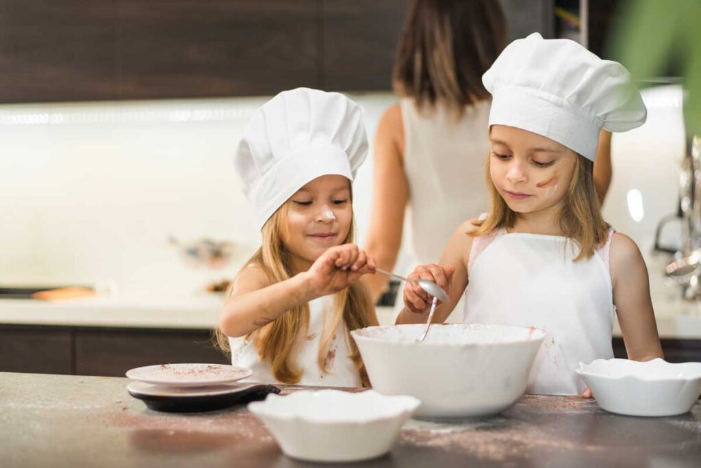 Dwie dziewczynki z czapkami kucharskimi na głowach mieszają w kuchni składniki na ciasto