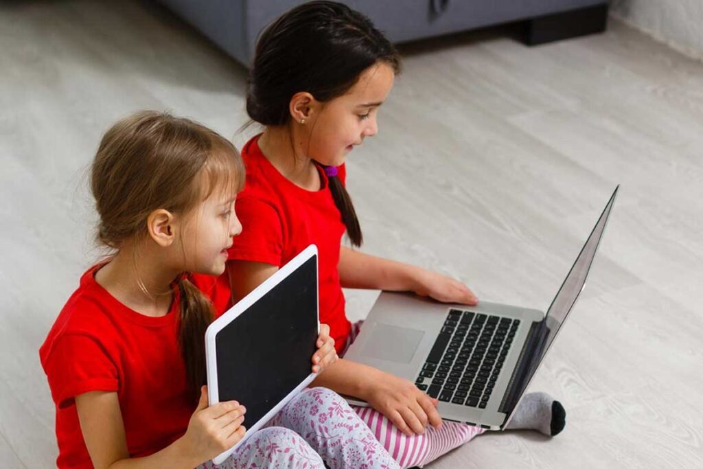 dwie dziewczynki siedzą na podłodze trzymając laptop i tablet