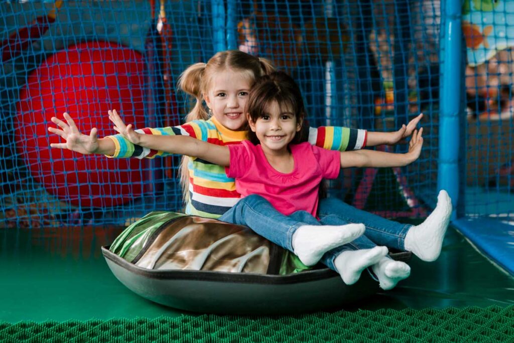 dwie radosne dziewczynki bawią się w sali zabaw zjeżdżając na oponie