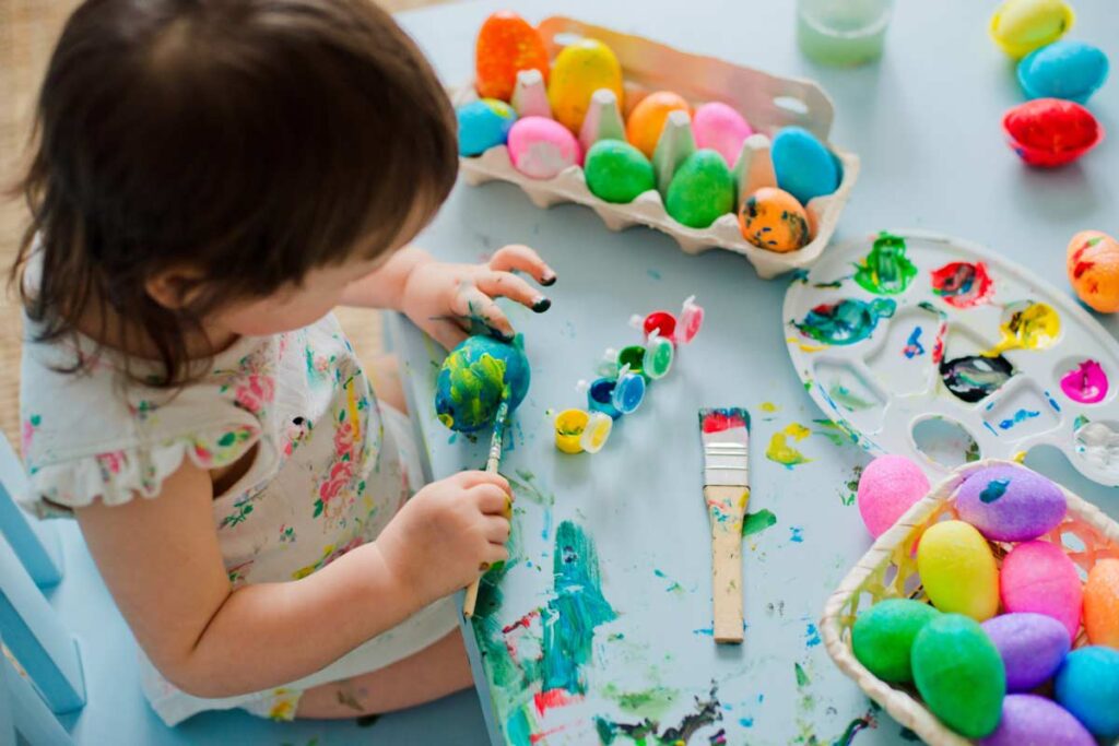 mała dziewczynka maluje wielkanocne jajka farbami na ubrudzonym stole